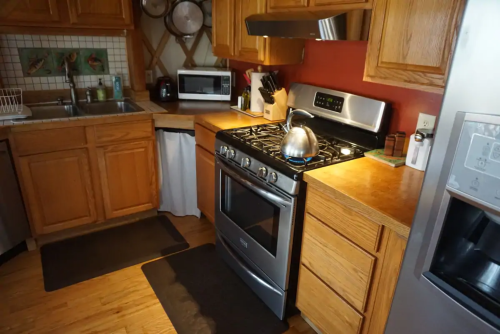 A cozy kitchen featuring wooden cabinets, a gas stove with a kettle, a microwave, and a sink with a dish rack.