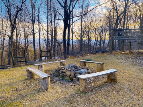 A serene outdoor fire pit area with wooden benches, surrounded by trees and a sunset sky in the background.