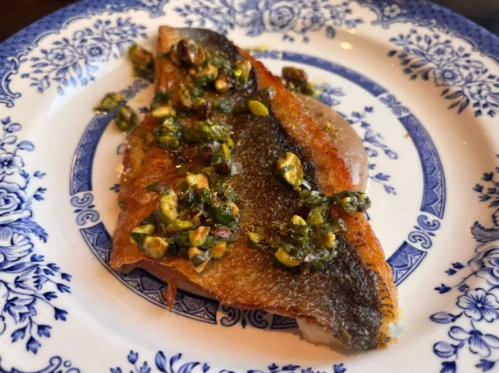 A piece of fish topped with a green herb and nut sauce, served on a decorative blue and white plate.