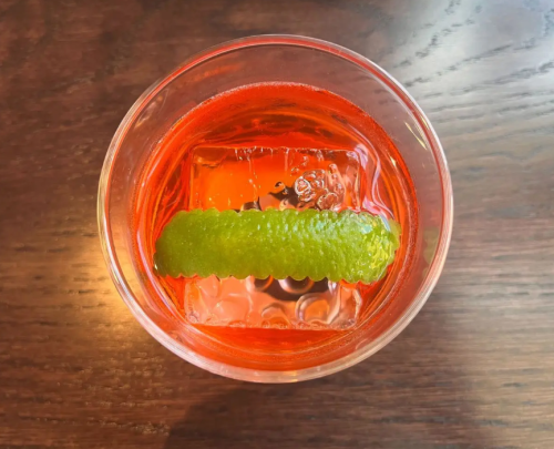 A close-up of a vibrant red cocktail with a lime twist on top, served over ice in a clear glass.