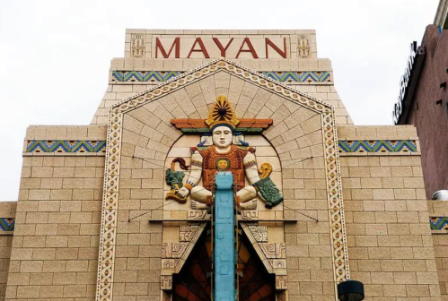 Art Deco-style building facade with "MAYAN" sign and intricate stone carvings, featuring a central figure and decorative elements.