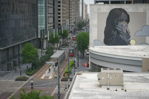 A city street scene featuring a large mural of a woman with a mask, surrounded by modern buildings and greenery.