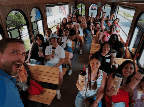 A group of people on a trolley, smiling and holding drinks, enjoying a fun outing together.
