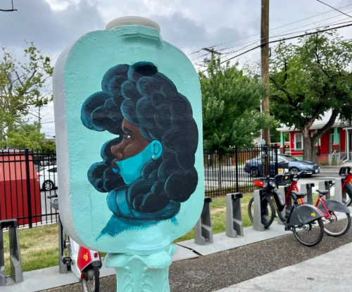 A mural of a woman with curly hair and a mask, painted on a utility box in an urban setting.