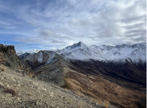 A rugged mountain landscape with snow-capped peaks under a cloudy sky, featuring rocky terrain and sparse vegetation.