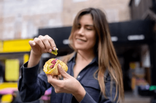 A woman smiles as she adds toppings to a taco outdoors, with a food truck in the background.