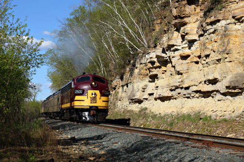 A vintage train travels along a rocky track surrounded by greenery and steep cliffs under a clear blue sky.
