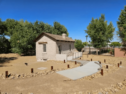 A small, historic stone house surrounded by trees and a gravel path in a sunny outdoor setting.