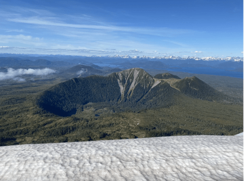 A panoramic view of a volcanic crater surrounded by lush forests and distant snow-capped mountains under a clear blue sky.