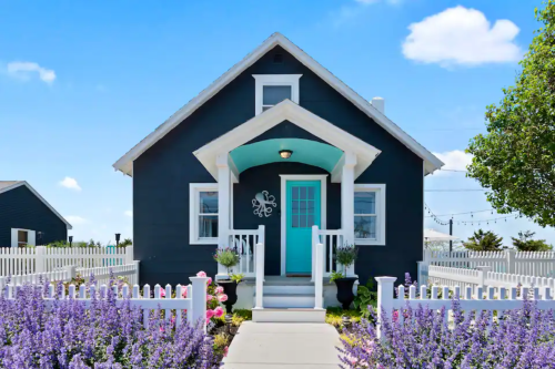 Charming black house with a turquoise door, surrounded by colorful flowers and a white picket fence under a blue sky.