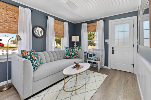 Cozy living room with a gray sofa, colorful pillows, a coffee table, and natural light from large windows.