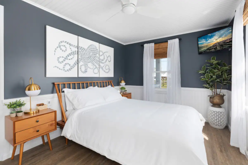 A modern bedroom with a white bed, wooden furniture, octopus artwork, and a TV on the wall, featuring light curtains.