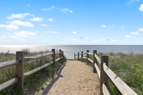 A sandy path leads to a calm sea, framed by wooden fences and grass under a bright blue sky.
