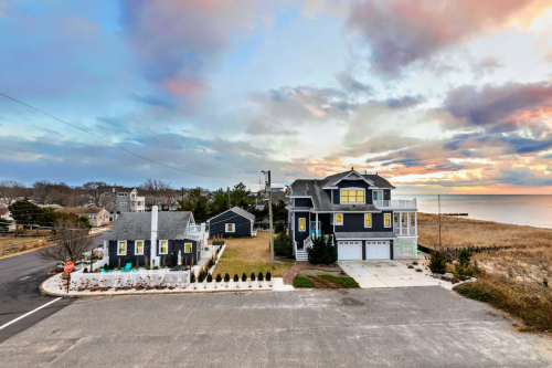 A coastal scene featuring two houses near the shore, with a colorful sunset and cloudy sky in the background.