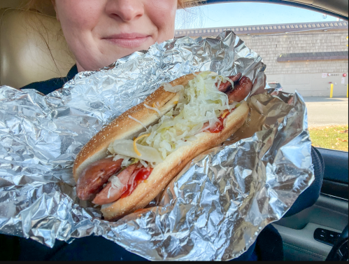 A person holding a hot dog with toppings, wrapped in foil, sitting in a car.