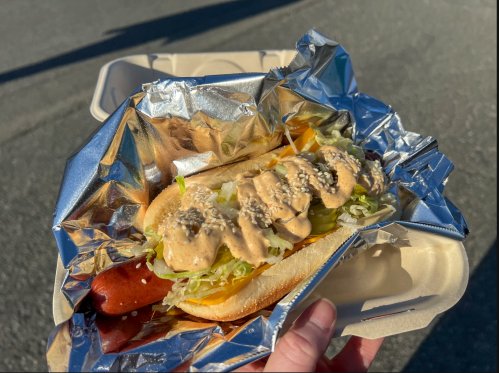 A hand holding a hot dog topped with lettuce, pickles, and a creamy sauce, wrapped in foil and placed in a takeout container.