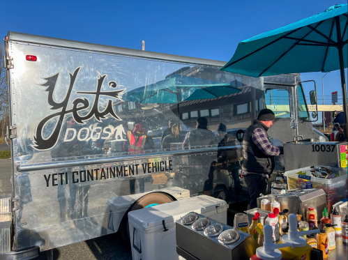 A shiny food truck named "Yeti Dogs" with a vendor preparing food under a blue umbrella on a sunny day.