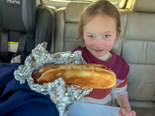 A young girl in a car holds up a foil-wrapped hot dog, smiling at the camera.