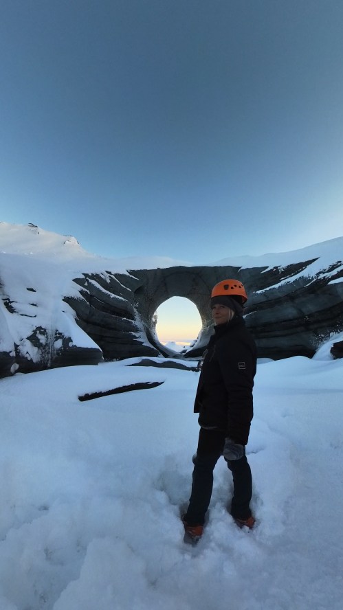 A person in an orange helmet stands on snow near a large, circular ice formation against a clear blue sky.