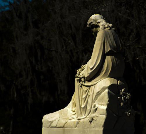 A serene statue of a woman sitting, holding flowers, surrounded by trees and soft shadows.