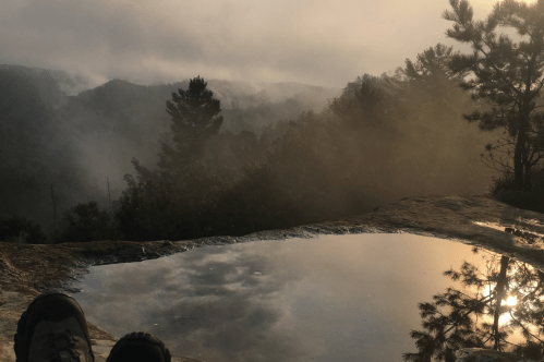 A serene landscape with misty mountains and a reflective puddle on a rock, viewed from a seated position.