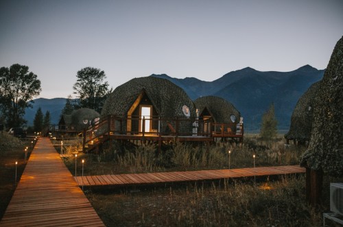 Cozy, dome-shaped cabins on wooden pathways, surrounded by nature at dusk with mountains in the background.