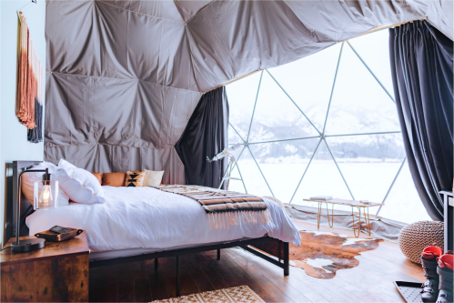 Cozy bedroom with a large window showcasing snowy mountains, featuring a bed, decorative items, and warm lighting.