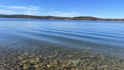 Calm lake water with clear visibility of rocks beneath, surrounded by hills under a blue sky.
