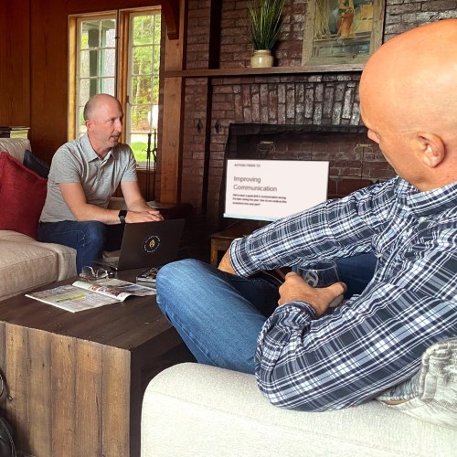 Two men engaged in a discussion in a cozy living room, with a laptop and presentation on improving communication.