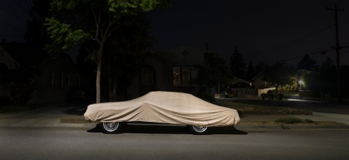 A covered car parked on a quiet street at night, illuminated by nearby streetlights and house lights.