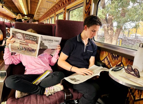 Two children sit on a train, one reading a newspaper while the other hides behind a newspaper, both focused on their reading.