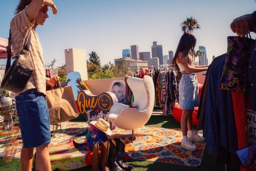 A vibrant outdoor market scene with people browsing colorful clothing and accessories against a city skyline backdrop.
