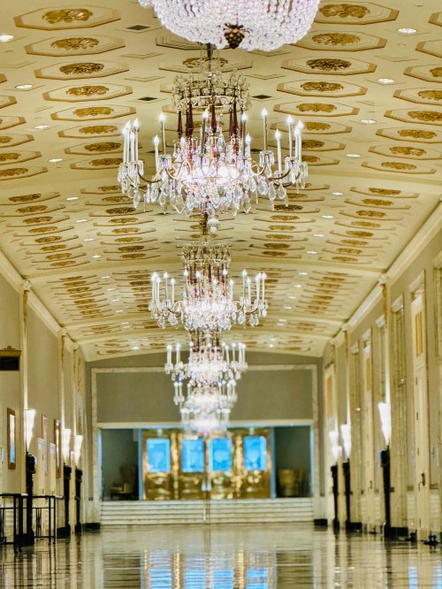 A grand hallway with ornate chandeliers, elegant ceiling designs, and polished floors reflecting the light.