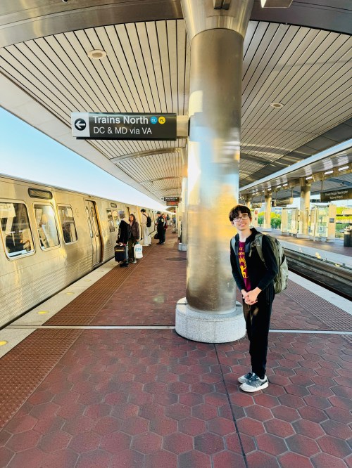 A young person stands at a train station platform with a train and passengers in the background, under a directional sign.