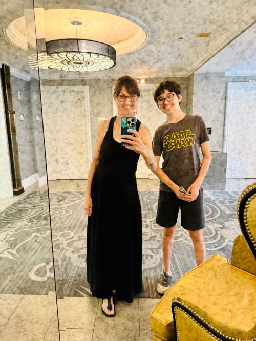A woman in a black dress and a boy in a Star Wars t-shirt pose for a selfie in a mirrored hallway.