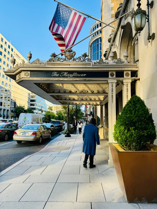 A man in a blue coat walks past the Mayflower Hotel, with an American flag waving above the entrance.