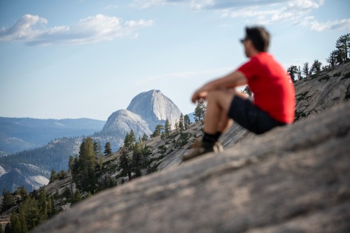 A person in a red shirt sits on a rocky surface, gazing at a distant mountain under a blue sky.