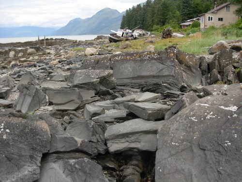 Rocky shoreline with engraved stones, a house in the background, and mountains under a cloudy sky.