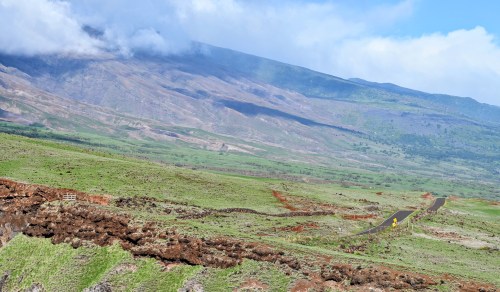 A winding road through lush green fields and rocky terrain, with mountains and clouds in the background.