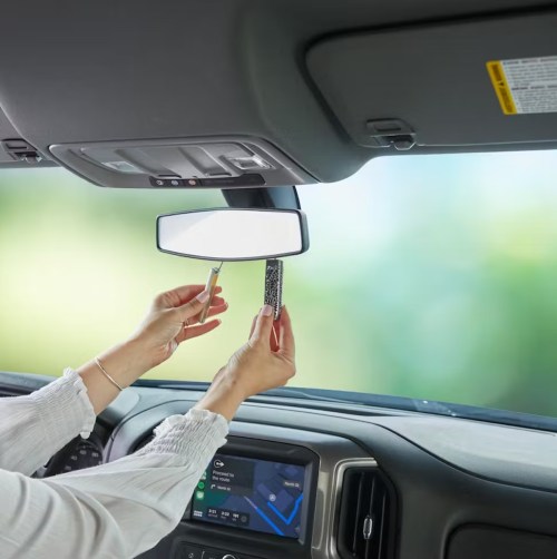 A person adjusts a rearview mirror in a car, holding a small object in one hand. The dashboard is visible below.