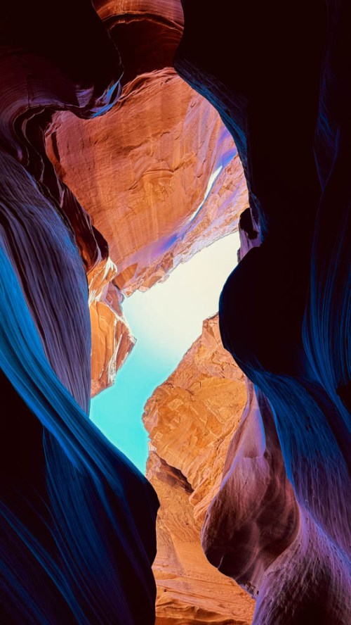 View looking up through a narrow canyon, showcasing vibrant orange and blue rock formations against a bright sky.