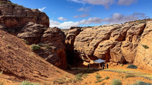 A rocky canyon with orange sandstone formations and a small shelter under a blue sky with scattered clouds.