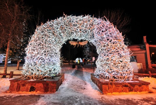 A large, illuminated arch made of antlers, surrounded by snow and trees, creating a festive winter scene at night.