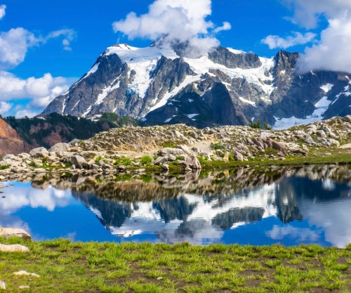 A stunning mountain landscape with snow-capped peaks reflected in a calm lake, surrounded by lush greenery and clouds.
