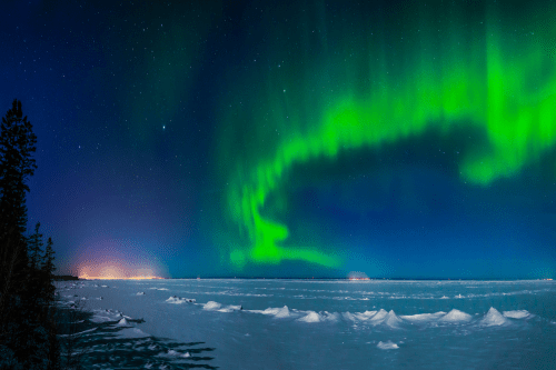 A panoramic view of the northern lights over a snowy landscape, with a starry sky and distant lights on the horizon.