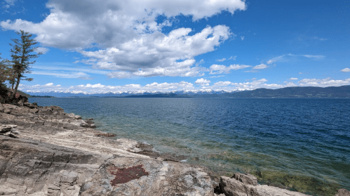 A scenic view of a lake with rocky shores, clear water, and mountains under a blue sky with fluffy clouds.