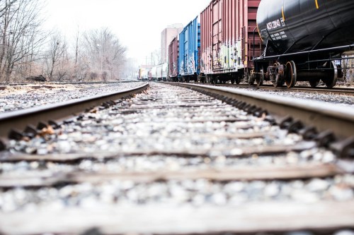 A low-angle view of train tracks lined with colorful freight cars, surrounded by bare trees and a cloudy sky.