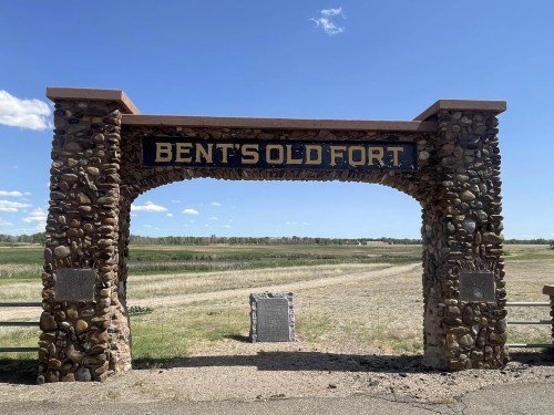 Archway sign for Bent's Old Fort, surrounded by open fields and a clear blue sky.