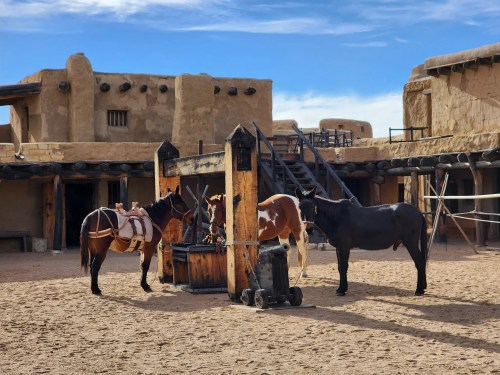 Three horses gather around a wooden water trough in a rustic, desert-like setting with adobe buildings in the background.