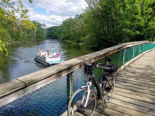 A bicycle rests on a wooden bridge overlooking a river, with a boat carrying people in the background.
